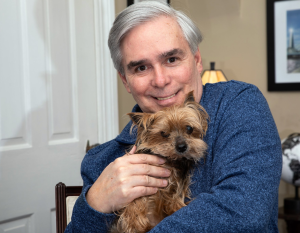 A photograph of a white man with gray hair wearing a blue sweater and holding a very small brown dog, some kind of terrier. Man and dog both look at the camera. The man is smiling at the camera. They are seated in what could be the living room of a well-decorated house, though its hard to be sure.